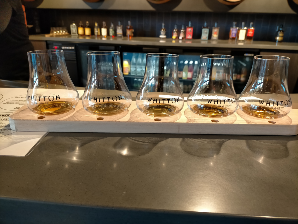 Five whisky glasses are lined up on a wooden paddle. A bar is visible in the background.