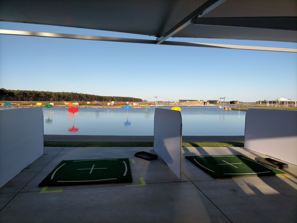 The image shows two golf tee-off mats facing a lake. The lake contains coloured funnels sticking out at regular intervals.