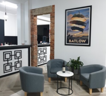 Three grey armchairs surround a small table with a bottle of wine on it. Behind them there is an artwork on the wall depicting the peel of an apple with the word 'BATLOW' below. On the left is a bar and an open doorway. 