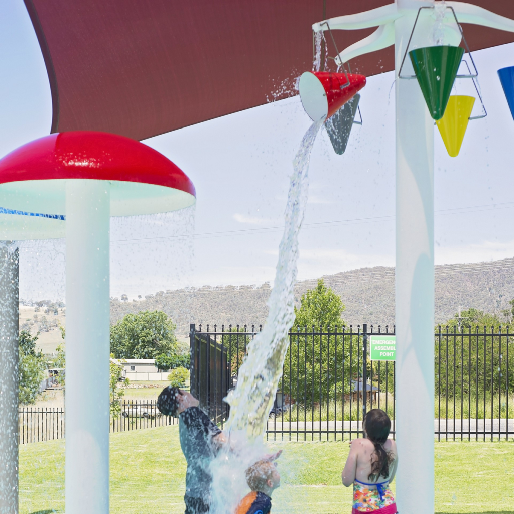 Image shows three children standing under a tower with coloured buckets suspended from it. One of the buckets is tipping water onto two of the children. 