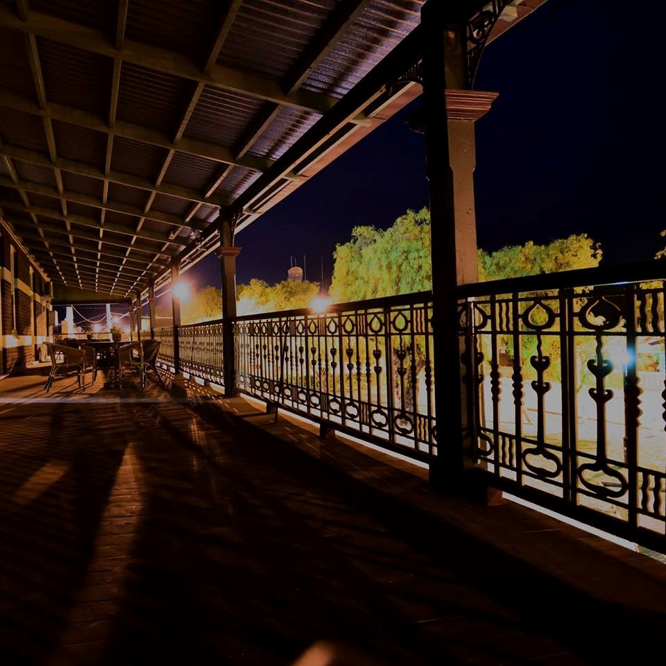 The image shows a wide verandah with decorative balustrade. It is nightime, and trees and streetlights are visible outside of the verandah.