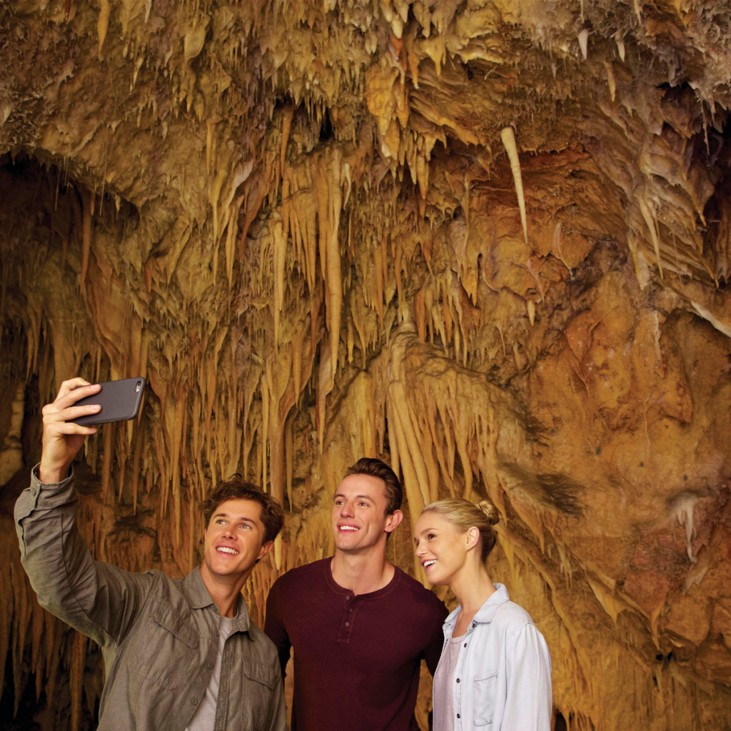 Image shows a group of two males and one female person, one male is holding a smartphone to take a selfie of the group. The group is inside a cave.