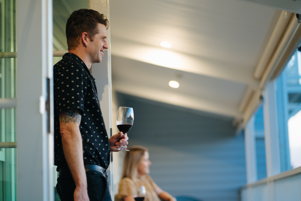 A man and woman are sitting on a balcony. The man is holding a glass of red wine. Another glass of red wine is in the background next to the woman.