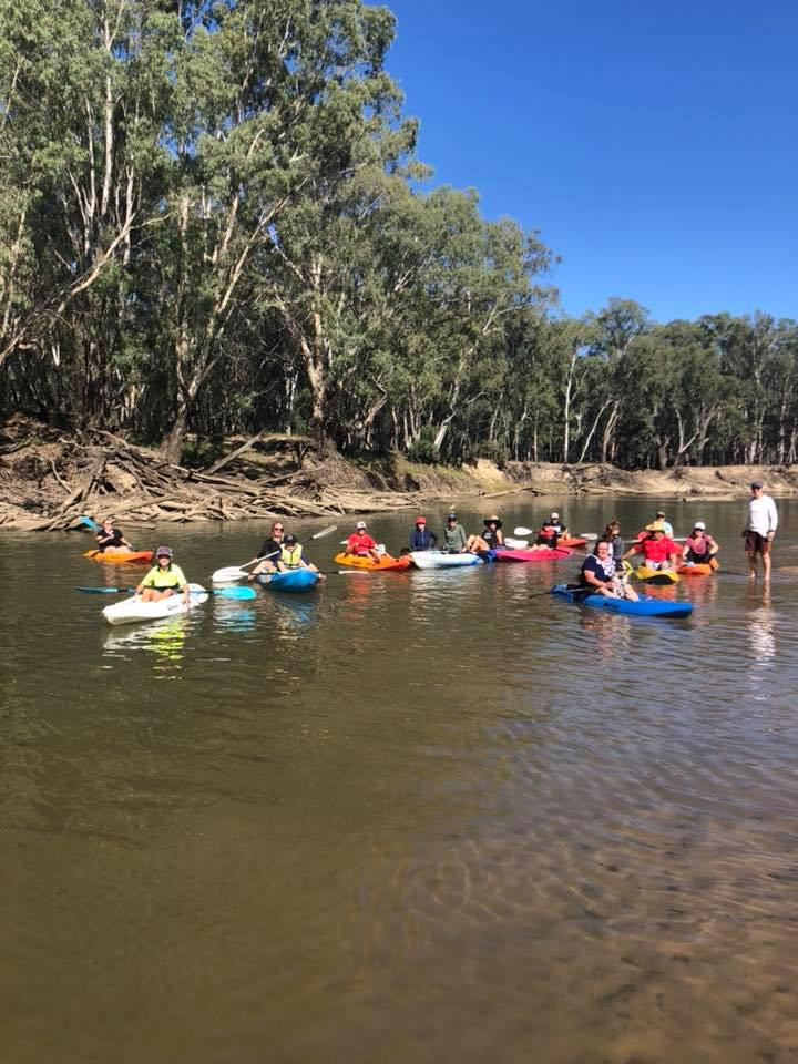 Image shows 15 people in kayaks on the murrumbidgee river and one man standing next to them.