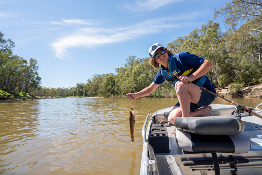 Image shows a man sitting on a boat in the murrumbidgee river. The man is holding a fishing rod in his left hand and a freshly-caught fish in his right hand. The man is smiling at the camera.