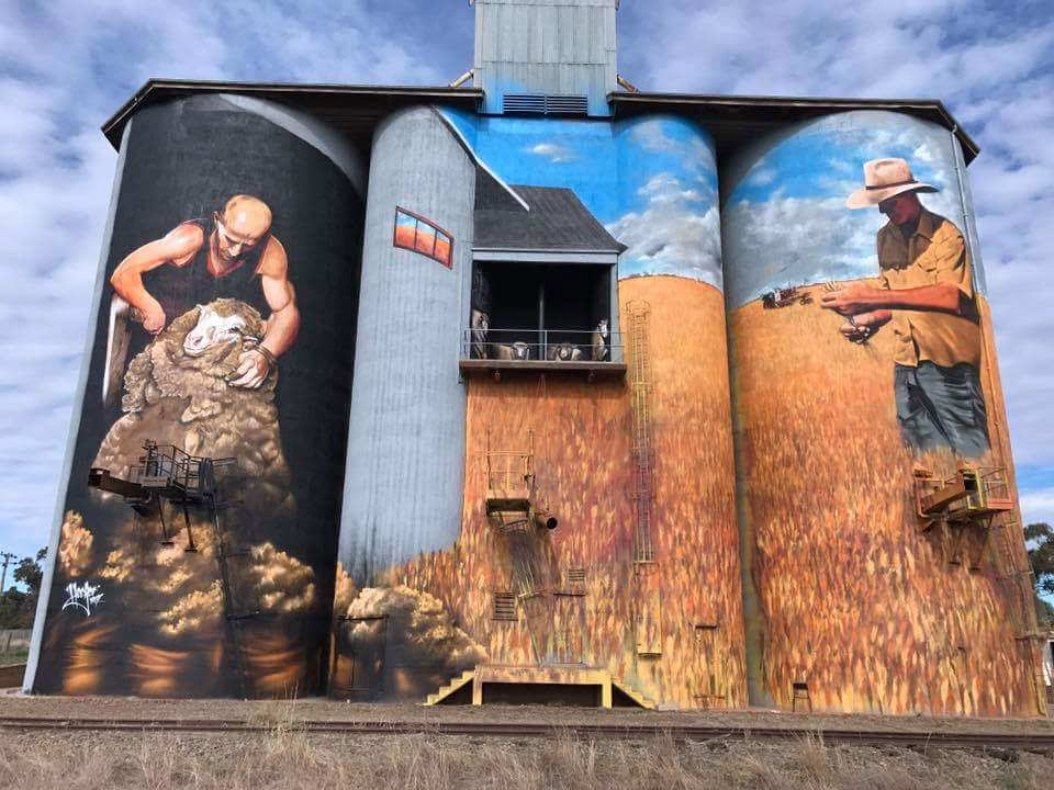 Three large silos stand tall against a blue sky mottled with clouds. The silos are painted with an image of a man shearing a sheet, and another standing in a field of grain.