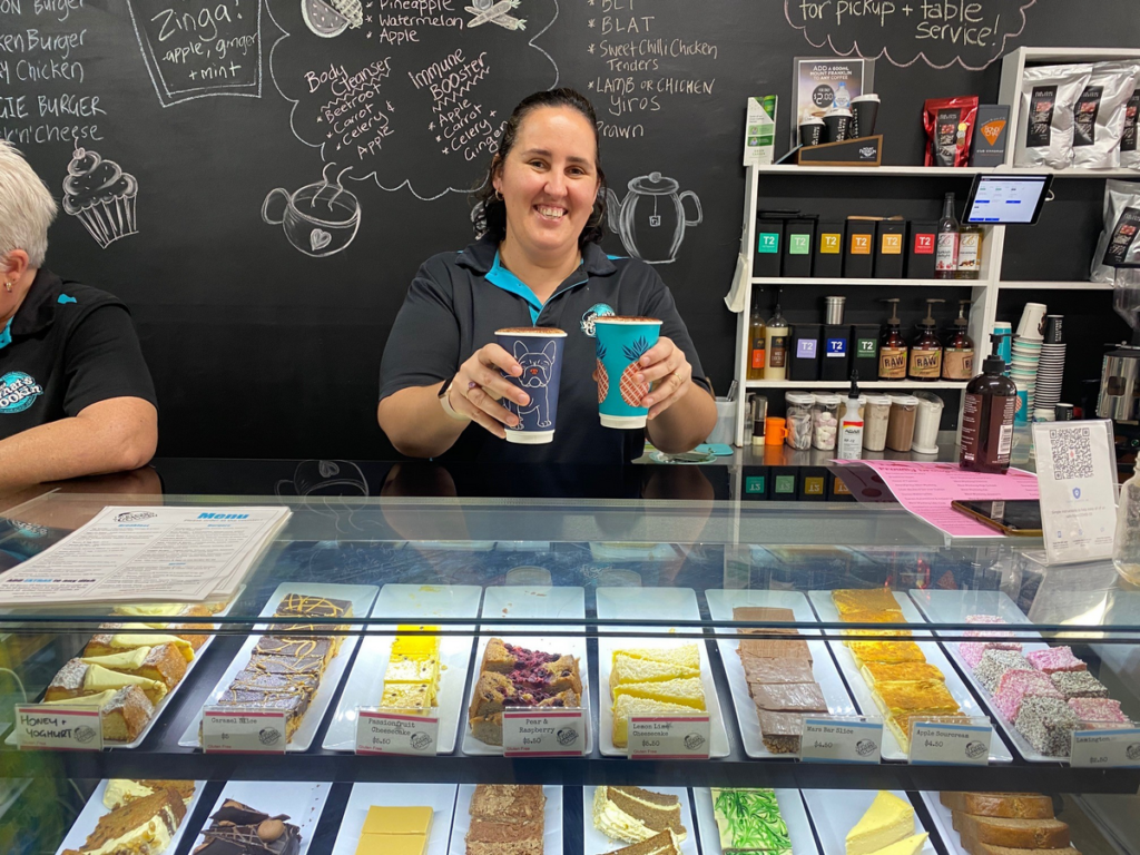 A woman is smiling and holding two large takeaway coffee cups. Behind her is a chalkboard showing menu options, and in front is a display of colourful cakes. 