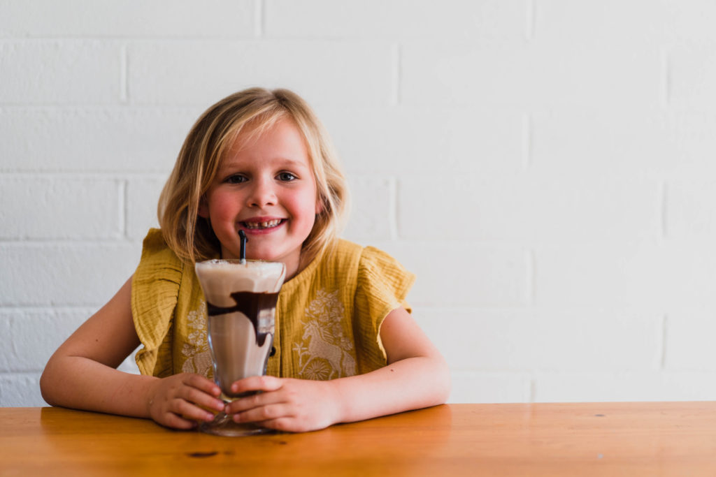 A young girl is smiling, sitting at a table and drinking a chocolate milkshake from a tall glass. 