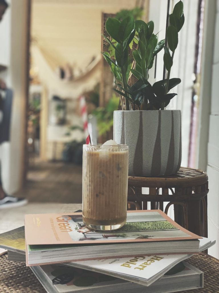 A tall glass containing a light brown drink and a red and white stripy straw sits on top of a pile of books and magazines. In the background there is a ceramic pot containing a green leafy plant, which is on top of a cane table.