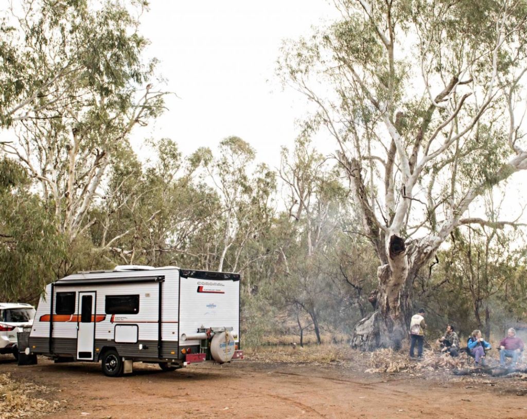 A Campervan is parked in an outback area. Four friends are gathered around a campfire. 