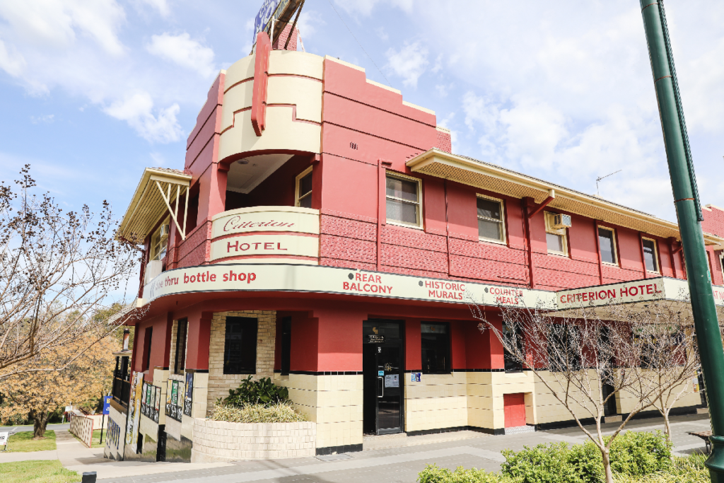 Image shows the external view of a large hotel in art deco style. The building is painted red and cream and a blue sky is visible above. 